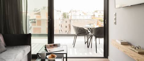 A modern living room with a sofa, a coffee table with a bowl and a magazine, a dining area with chairs and a table, and a large window with a view of buildings.