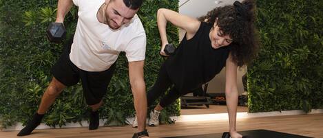 Two people exercising with dumbbells on a black mat in a room with a green wall.