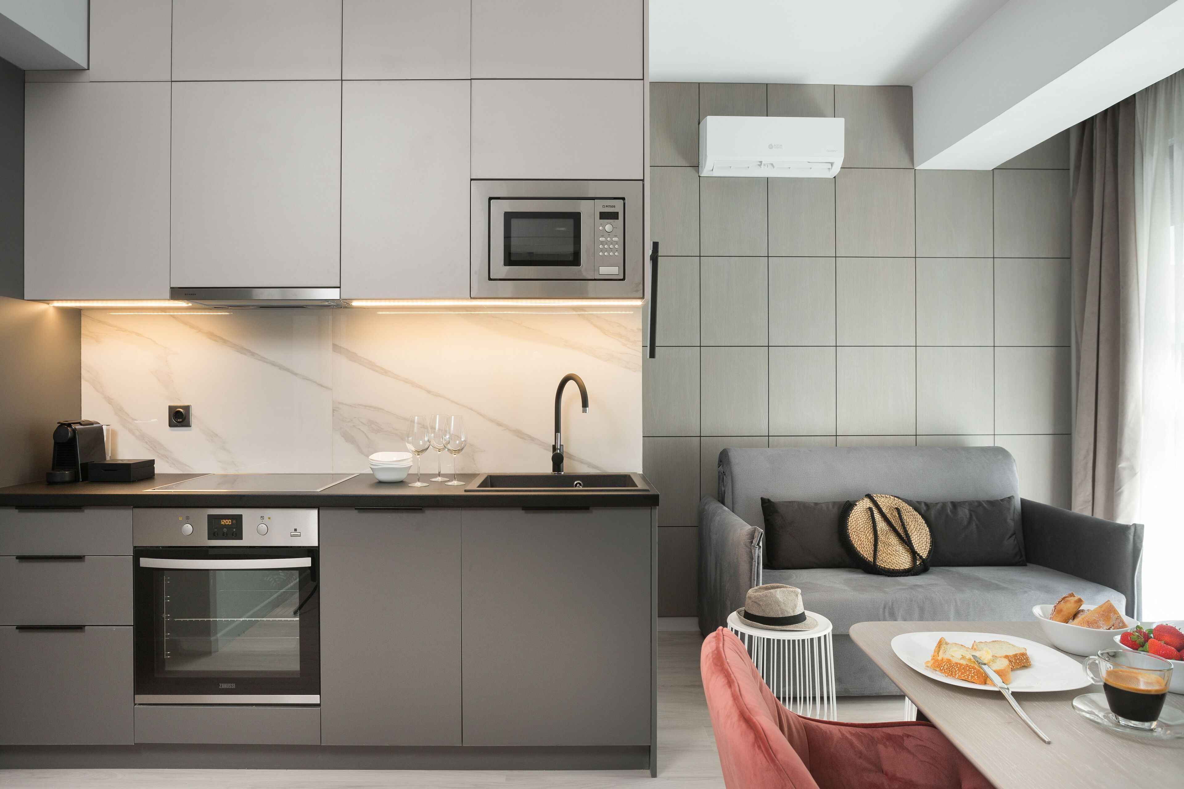A modern kitchen with a built-in oven, microwave, and sink. There is a dining area with a table set for two, a sofa, and a small side table with a hat and a bowl of fruit.