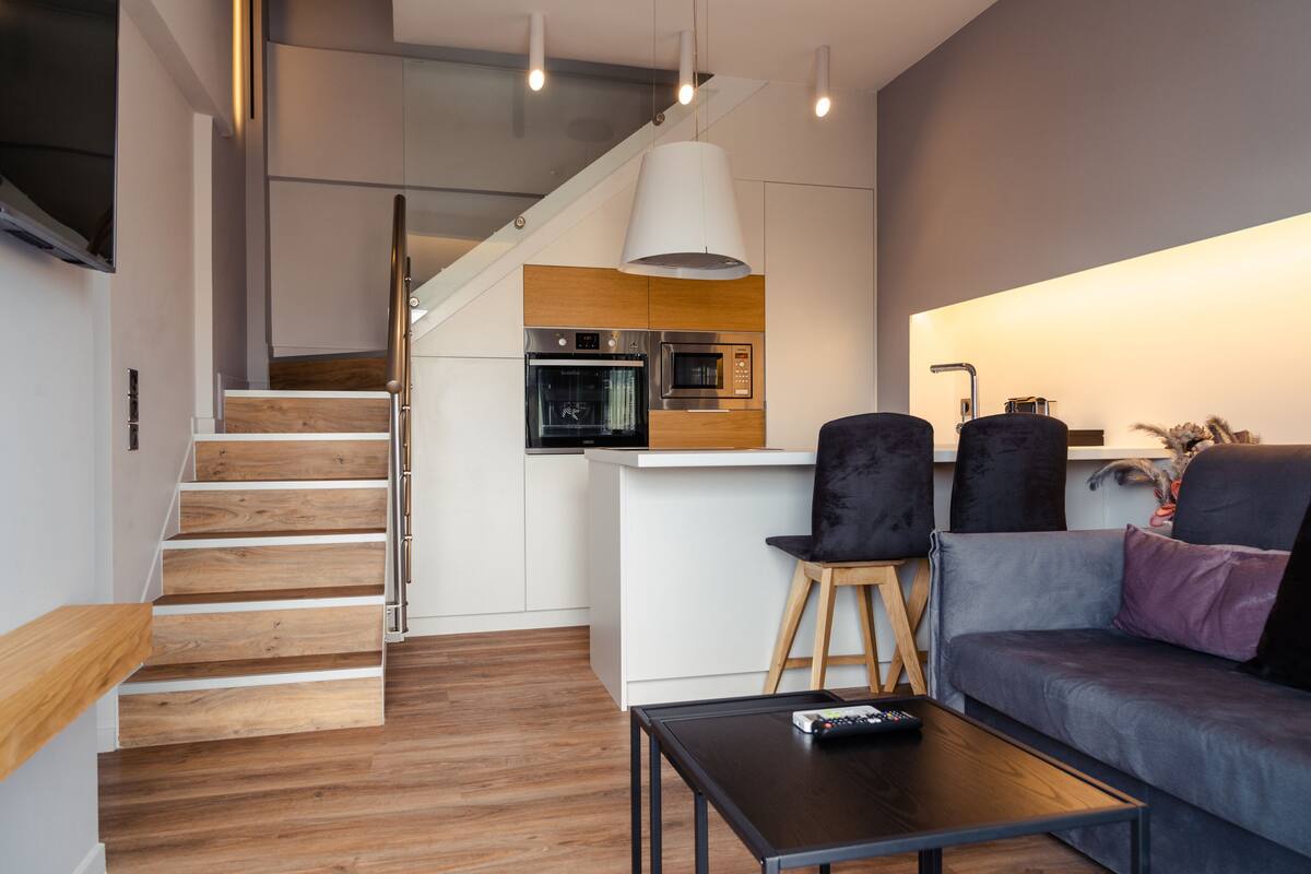 Une cuisine moderne équipée d’un four à micro-ondes, d’un four et de meubles en bois. Un escalier mène à un autre niveau. On y trouve également un canapé gris avec une télécommande posée sur une table, et un luminaire suspendu au-dessus de la cuisine.