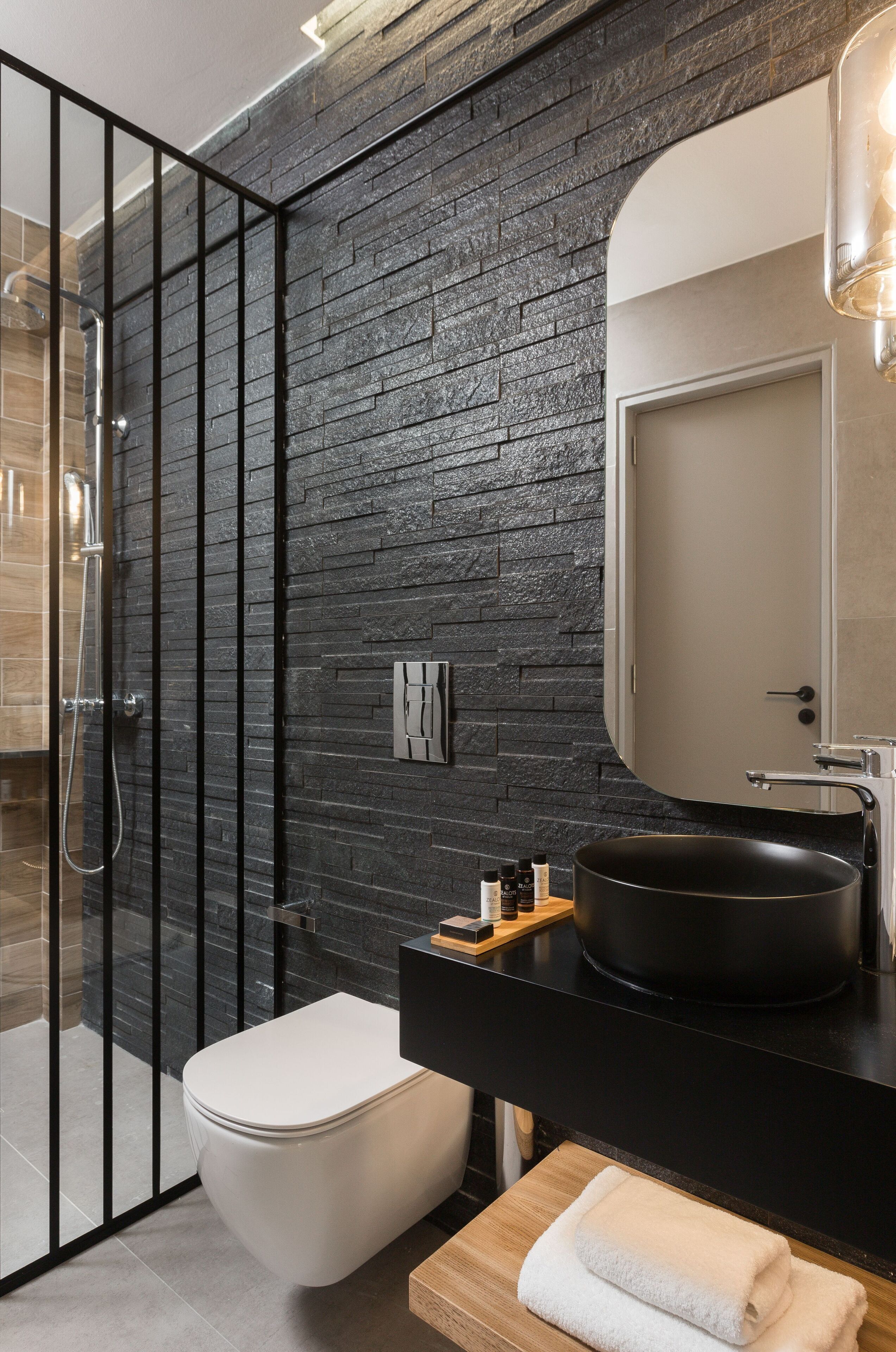 A modern bathroom with a black stone wall, a glass shower enclosure, a white toilet, and a black sink with a round basin.