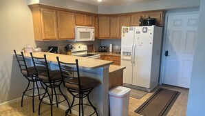 A kitchen with wooden cabinets, a white refrigerator, a microwave, a stove, and a sink. There are three bar stools and a trash can. - Can't Beat the View! (Camdenton)