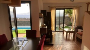 Ein modernes Wohnzimmer mit einem Glastisch, roten Kissen und Blick auf einen Pool und Grünflächen.