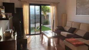 Ein Wohnzimmer mit einer Couch, einem Couchtisch und einem Fernseher auf einem Schrank. Eine Glastür führt auf einen Balkon mit Blick auf einen Garten und eine Palme.