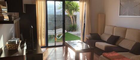 A living room with a sofa, a coffee table, and a TV on a stand. There is a sliding glass door leading to a balcony with a view of a garden and a palm tree.
