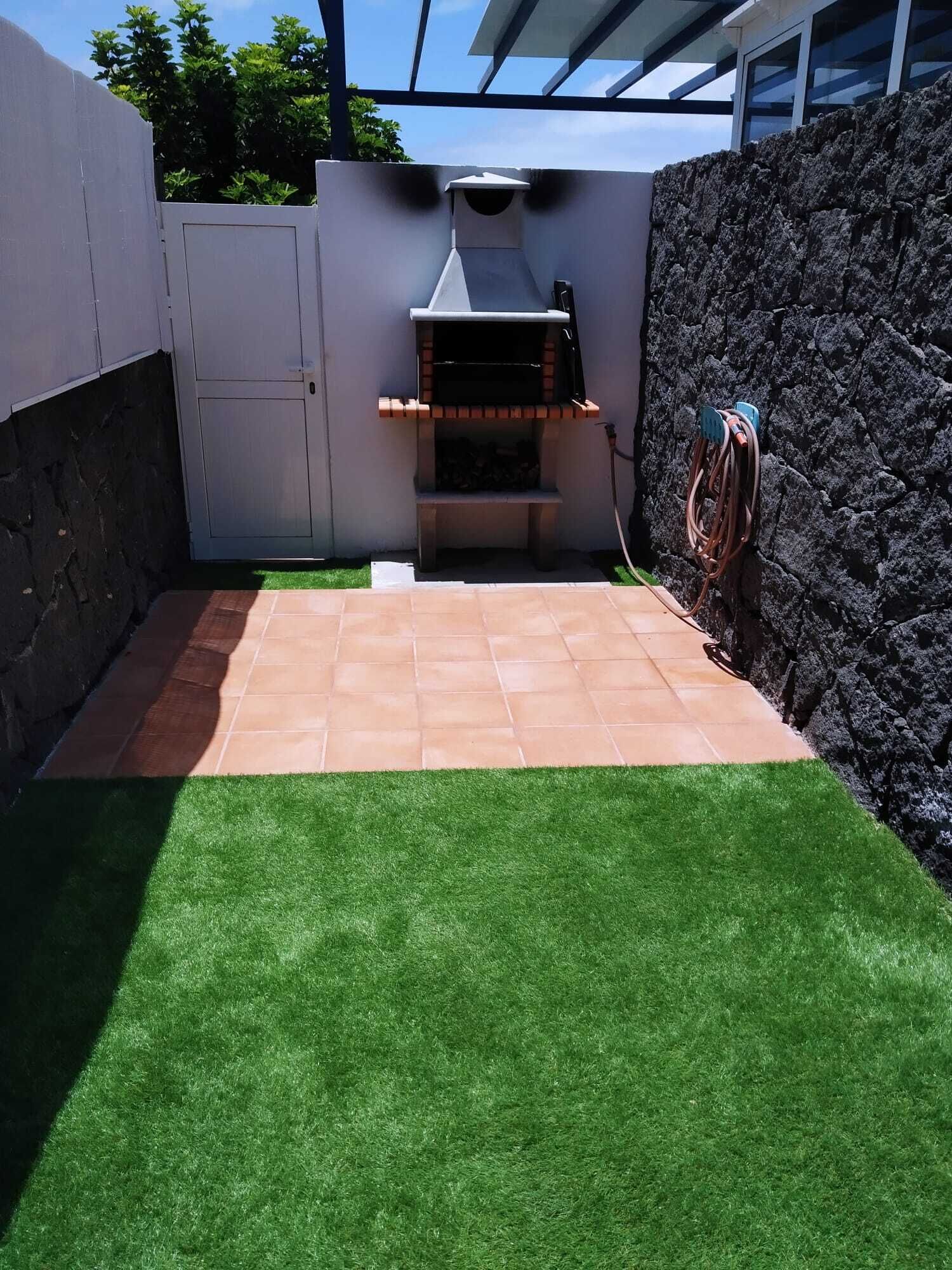 A backyard with a barbecue grill, artificial grass, a stone wall, and a white fence.