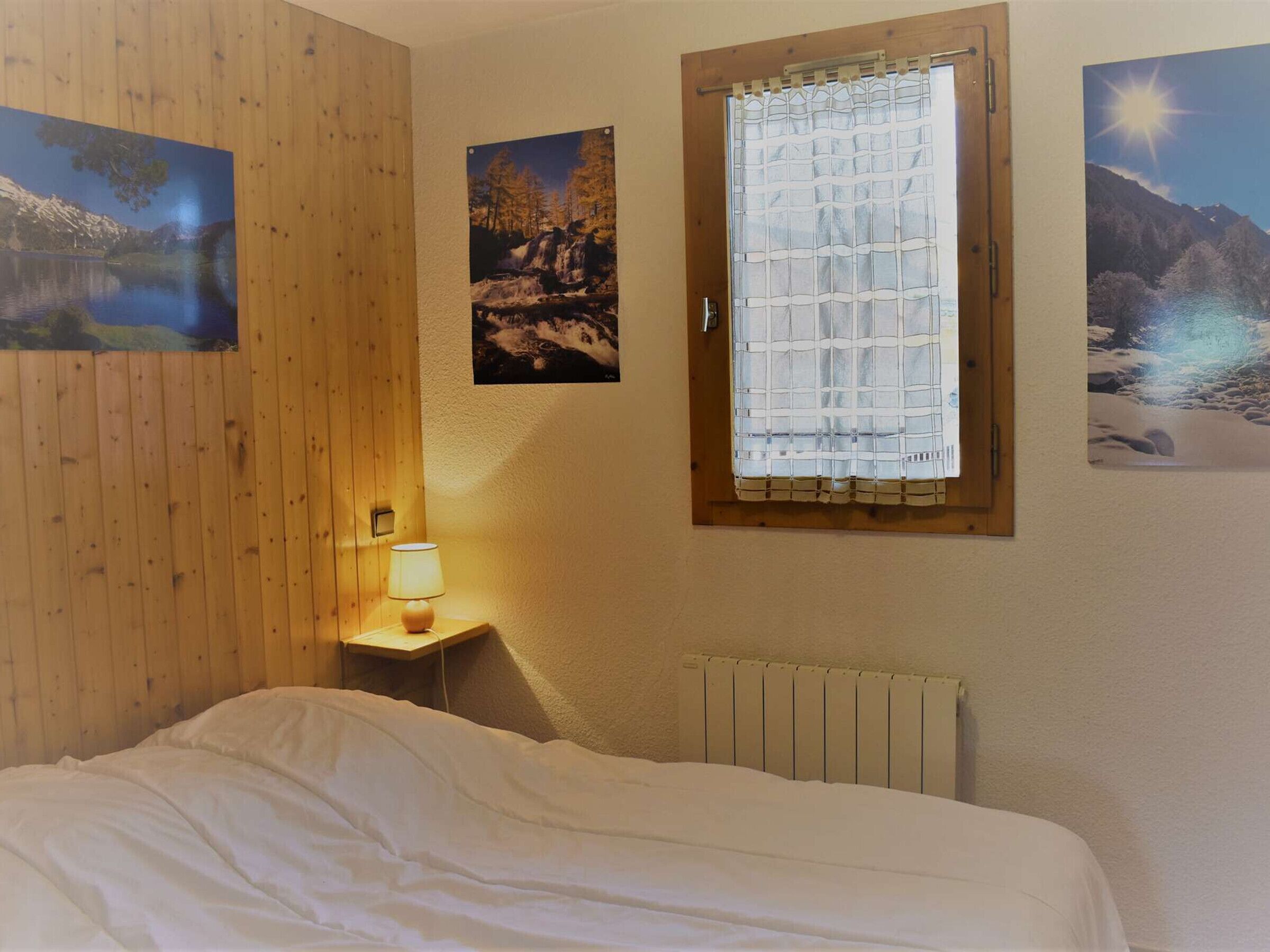 Une chambre avec un lambris en bois, une fenêtre avec un rideau, une lampe de chevet et trois tableaux encadrés accrochés au mur.