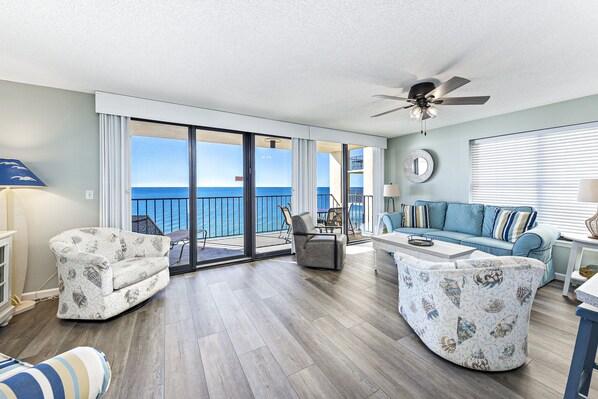 A spacious living room with a large sliding glass door leading to a balcony with a view of the ocean. There are two sofas, a coffee table, and a ceiling fan.