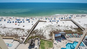A beach with numerous umbrellas and lounge chairs, a swimming pool, and a wooden walkway leading to the beach.