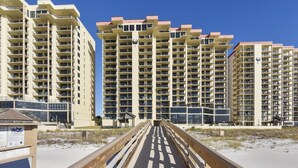 A beachfront hotel with multiple high-rise buildings, a wooden boardwalk leading to the beach, and a clear blue sky.