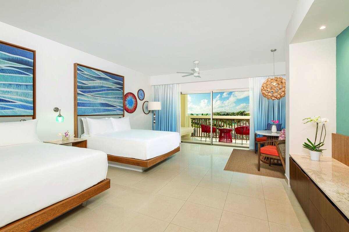 A hotel room with two beds, a balcony with red chairs, and a dining area with a table and chairs.