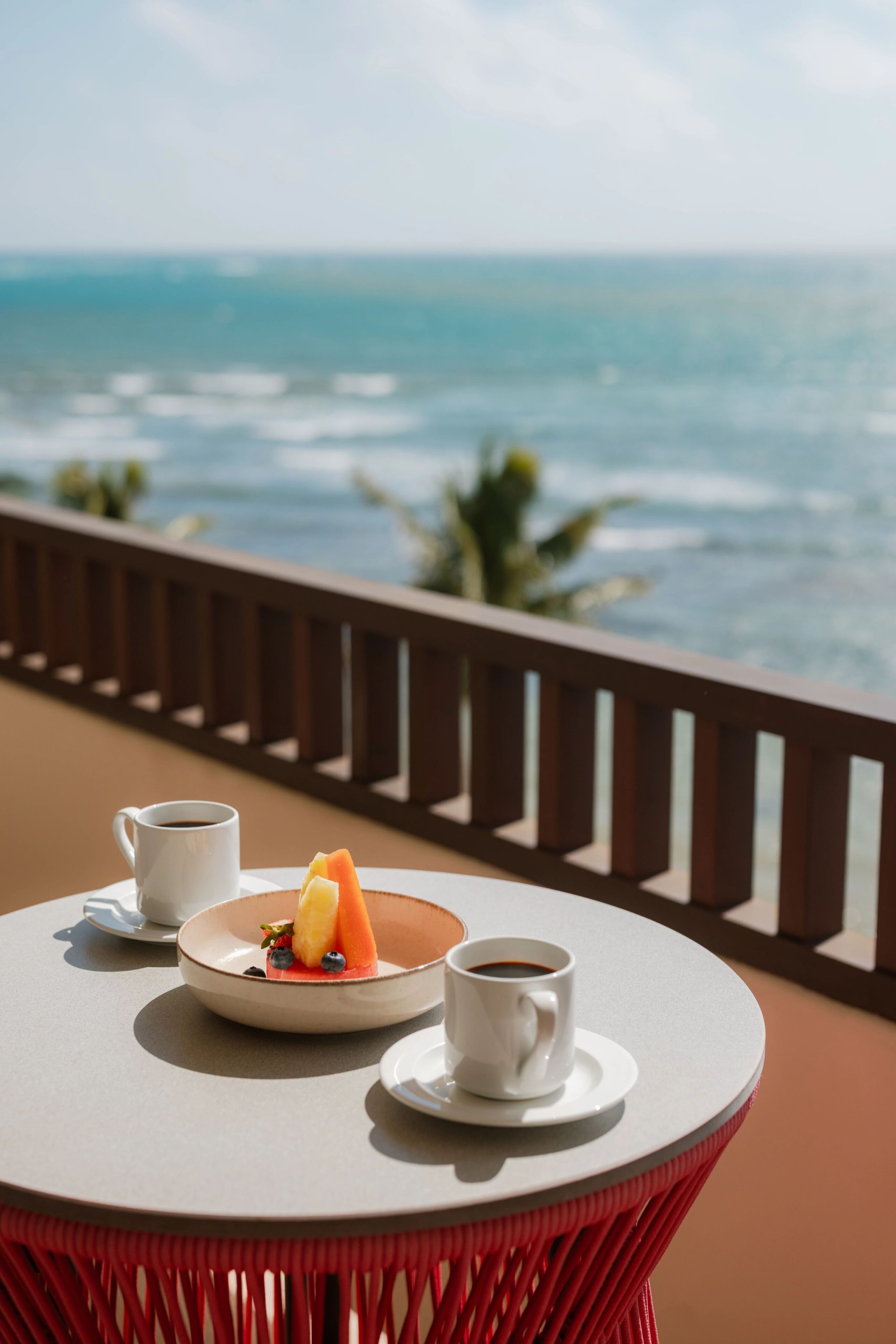 A round table with a white cup, a plate with fruit, and another white cup on a balcony overlooking the ocean.
