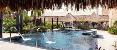 A swimming pool with a water feature, surrounded by thatched umbrellas and lush greenery.
