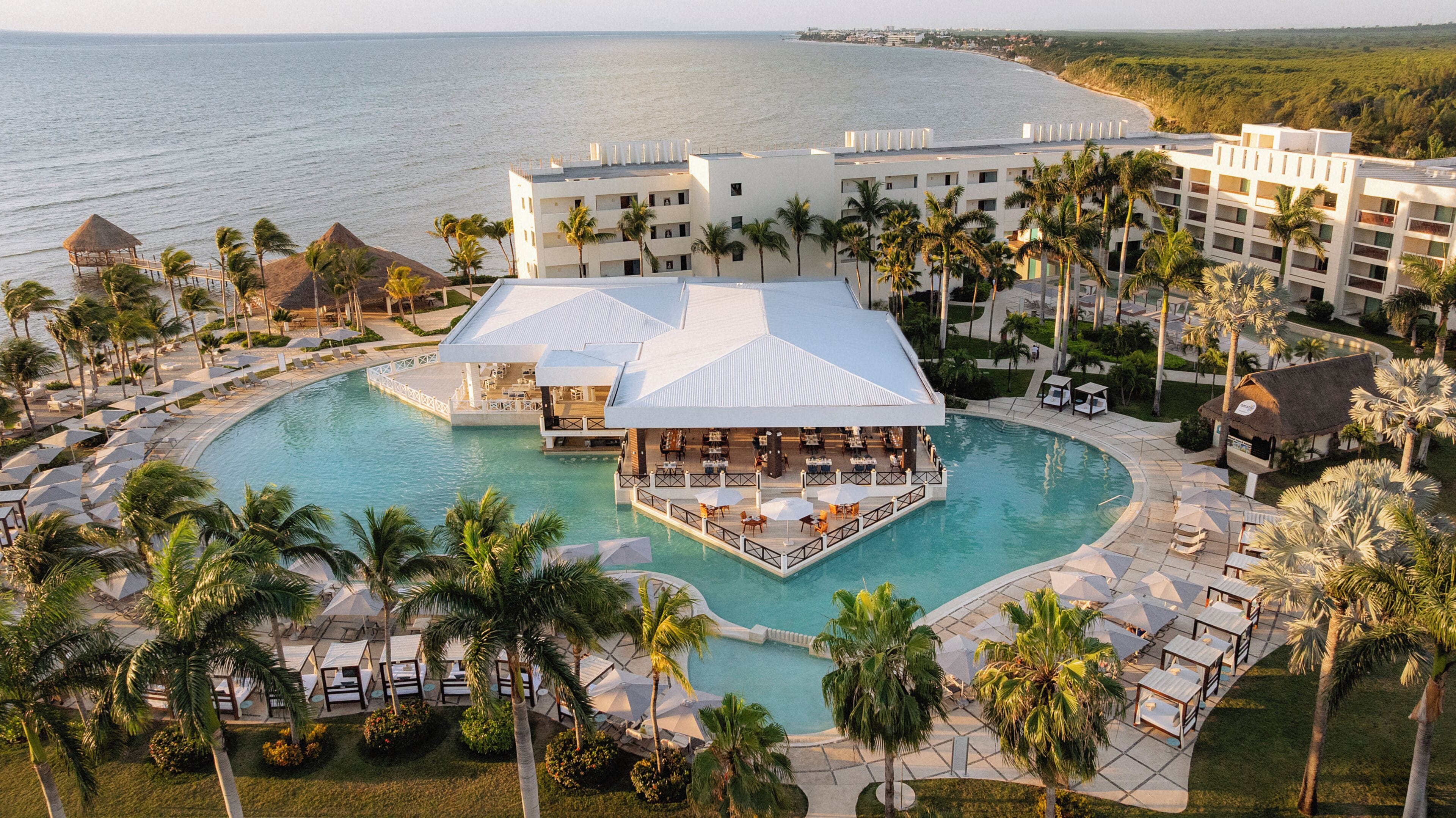 Aerial view of a curved pool beside resort buildings and the beach at Dreams Puerto Morelos Resort & Spa