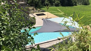 A pool area with a wooden deck, a shaded area, and a view of a green field and trees.