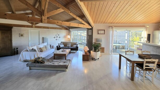 A spacious living area with a high ceiling, wooden beams, a sofa set, a dining table with chairs, and a view through large windows.