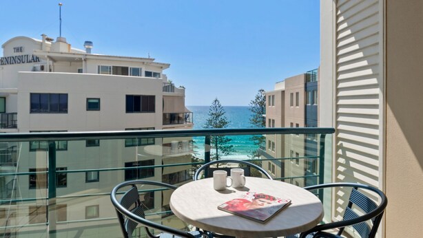 Un balcon avec une table, deux chaises et un magazine. La vue comprend un immeuble portant l’inscription « The Peninsula » et une étendue d’eau.