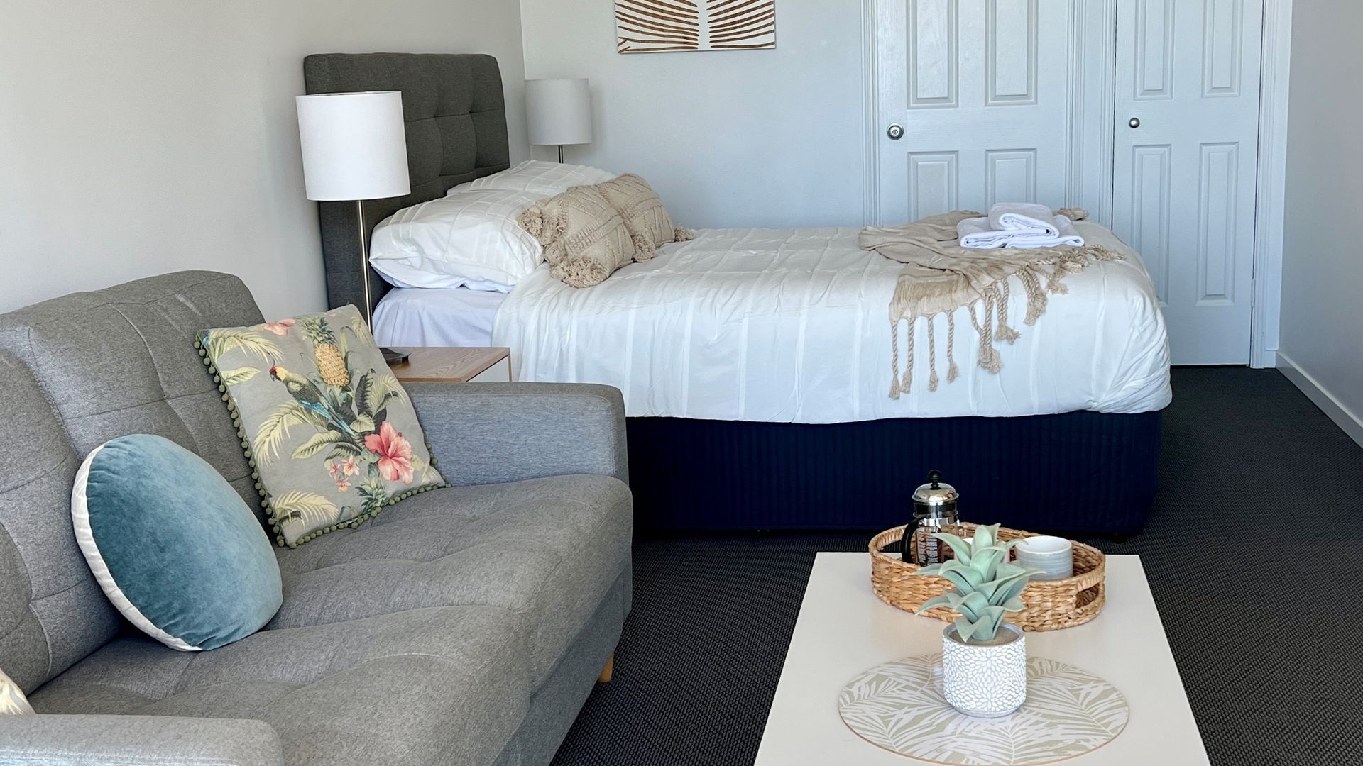 A hotel room with a grey sofa, a bed with white bedding, a lamp, a door, and a small table with a plant and a basket.