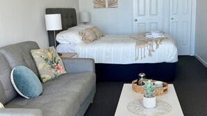 Une chambre d’hôtel avec un canapé gris, un lit avec une literie blanche, une lampe, une porte et une petite table sur laquelle se trouvent une plante et un panier.