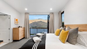 A modern hotel room with a large bed, a wooden dresser, and a view of mountains and a lake. - Oliver's Oasis | Luxury home | Spa Pool | BBQ | Fire (Queenstown)