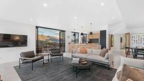 A modern living room with a large window offering a mountain view, a kitchen area with a dining table, and a cozy seating area with a sofa and chairs. - Oliver's Oasis | Luxury home | Spa Pool | BBQ | Fire (Queenstown)