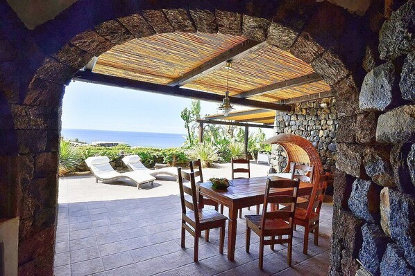 An outdoor dining area with stone walls, a thatched roof, and a view of the sea.