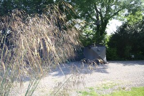 Zona de grava con pastos altos y una pequeña construcción, posiblemente un cobertizo, en un entorno similar a un parque.