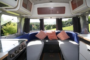 Interior compacto de una caravana, con kitchenette, zona de comedor y una litera con cojines.