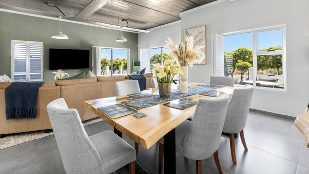 Una sala de estar moderna con una mesa de comedor de madera, sillas tapizadas en gris, un sofá color beige y un ventanal que ofrece vistas a zonas verdes.