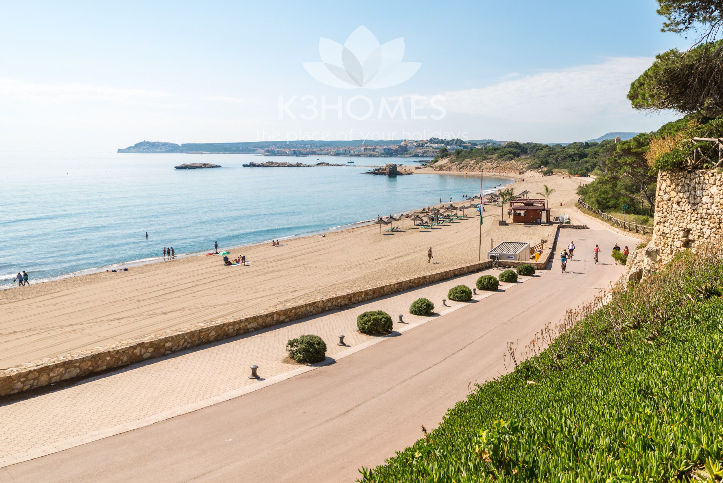 A coastal promenade with a stone pathway, beach, and a view of the sea and distant landforms.