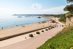 A coastal promenade with a stone pathway, beach, and a view of the sea and distant landforms.