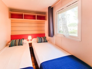 A hotel room with two single beds, a nightstand with a lamp, a window with a view of trees, and a red headboard.