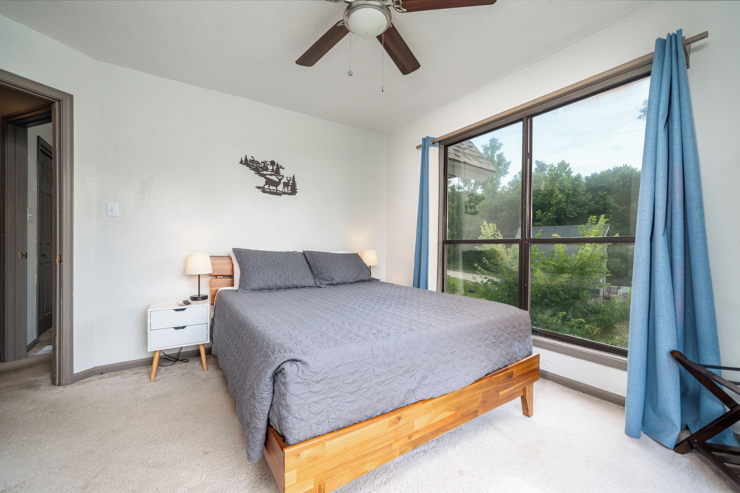 A bedroom with a bed, a nightstand, a ceiling fan, a window with curtains, and a view of greenery outside.