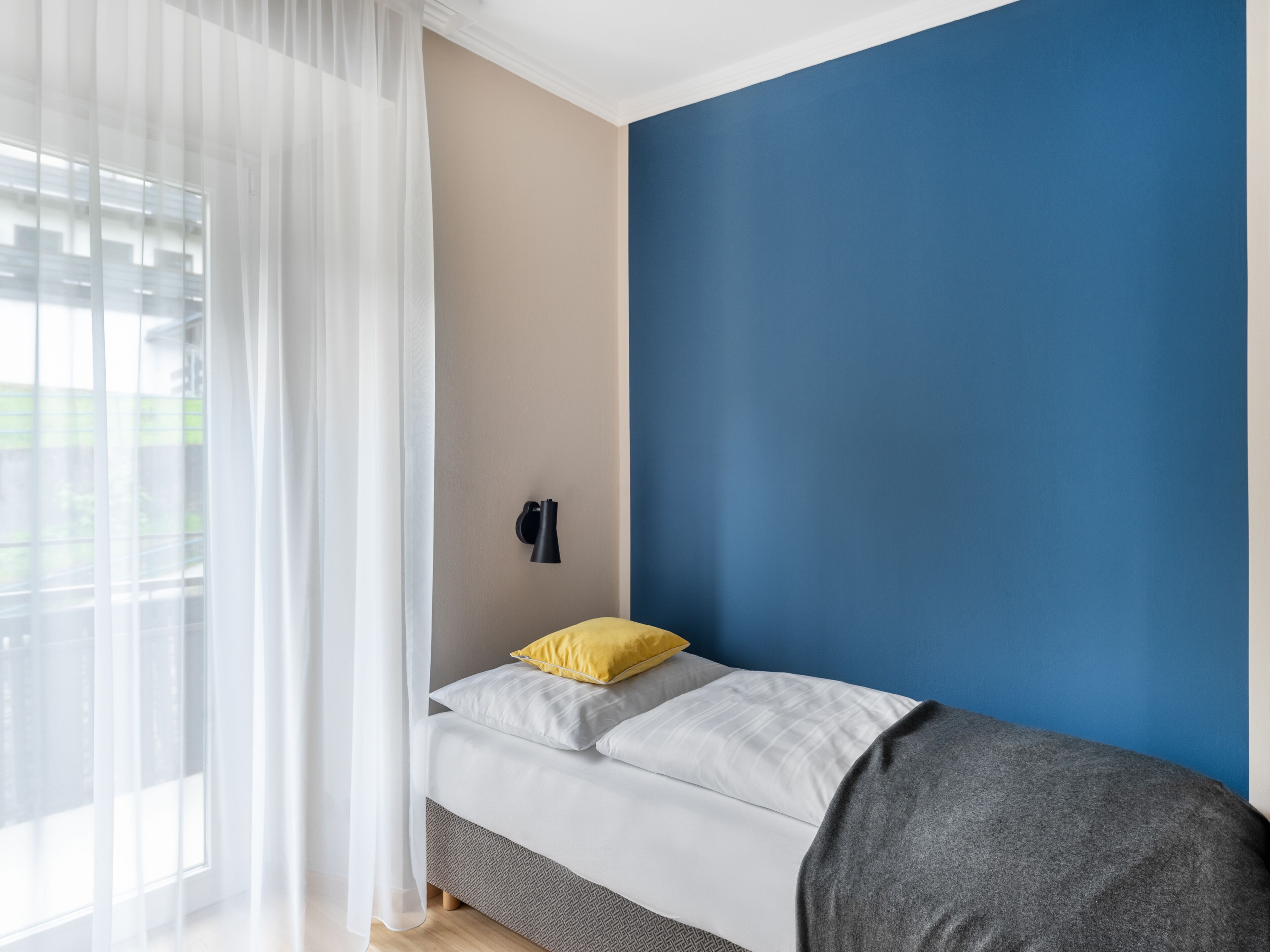 Un dormitorio con cama, ventana con cortinas translúcidas y pared pintada en dos colores.