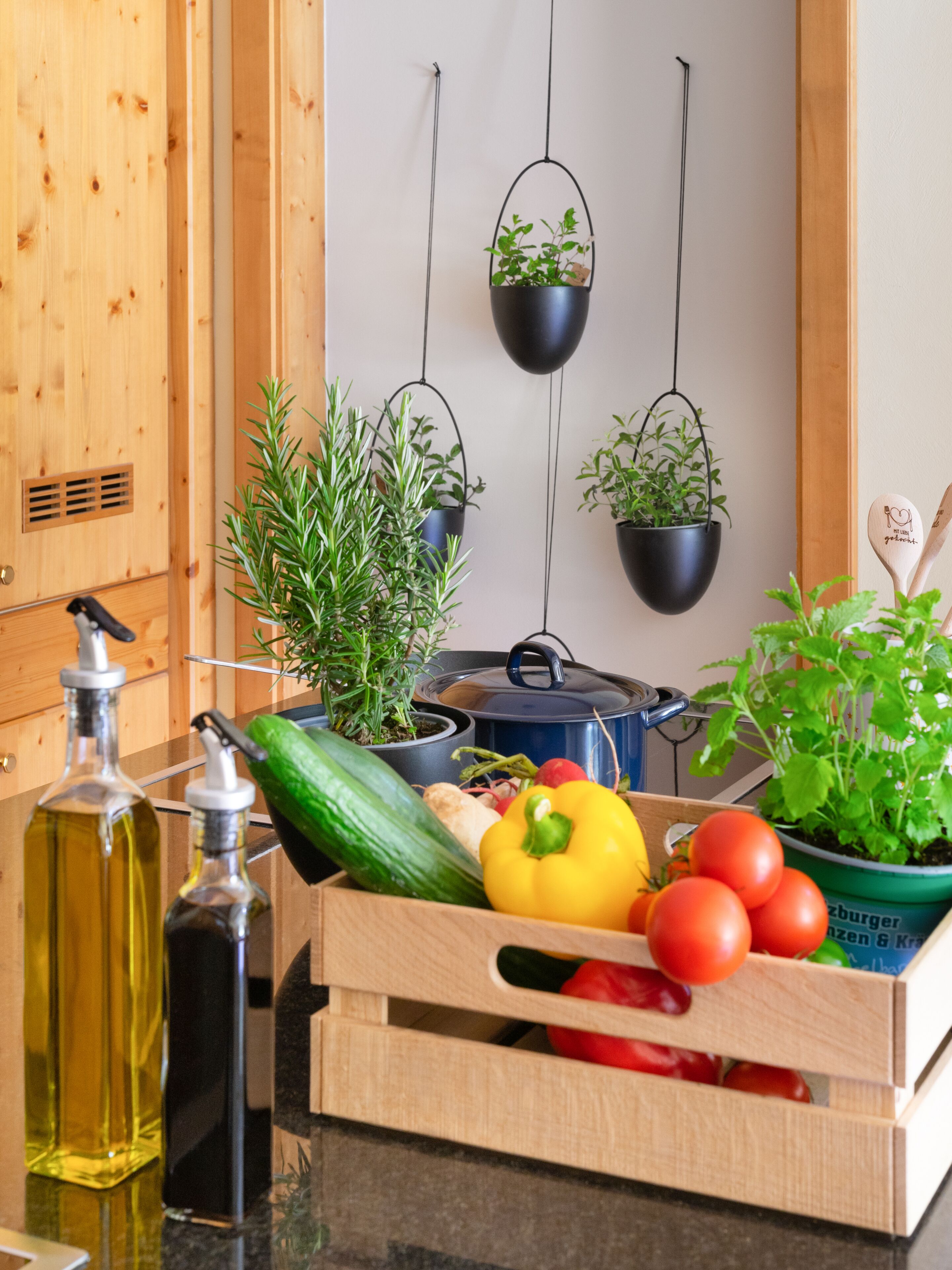 A kitchen with wooden cabinets, hanging plants, and a wooden crate filled with vegetables and herbs.