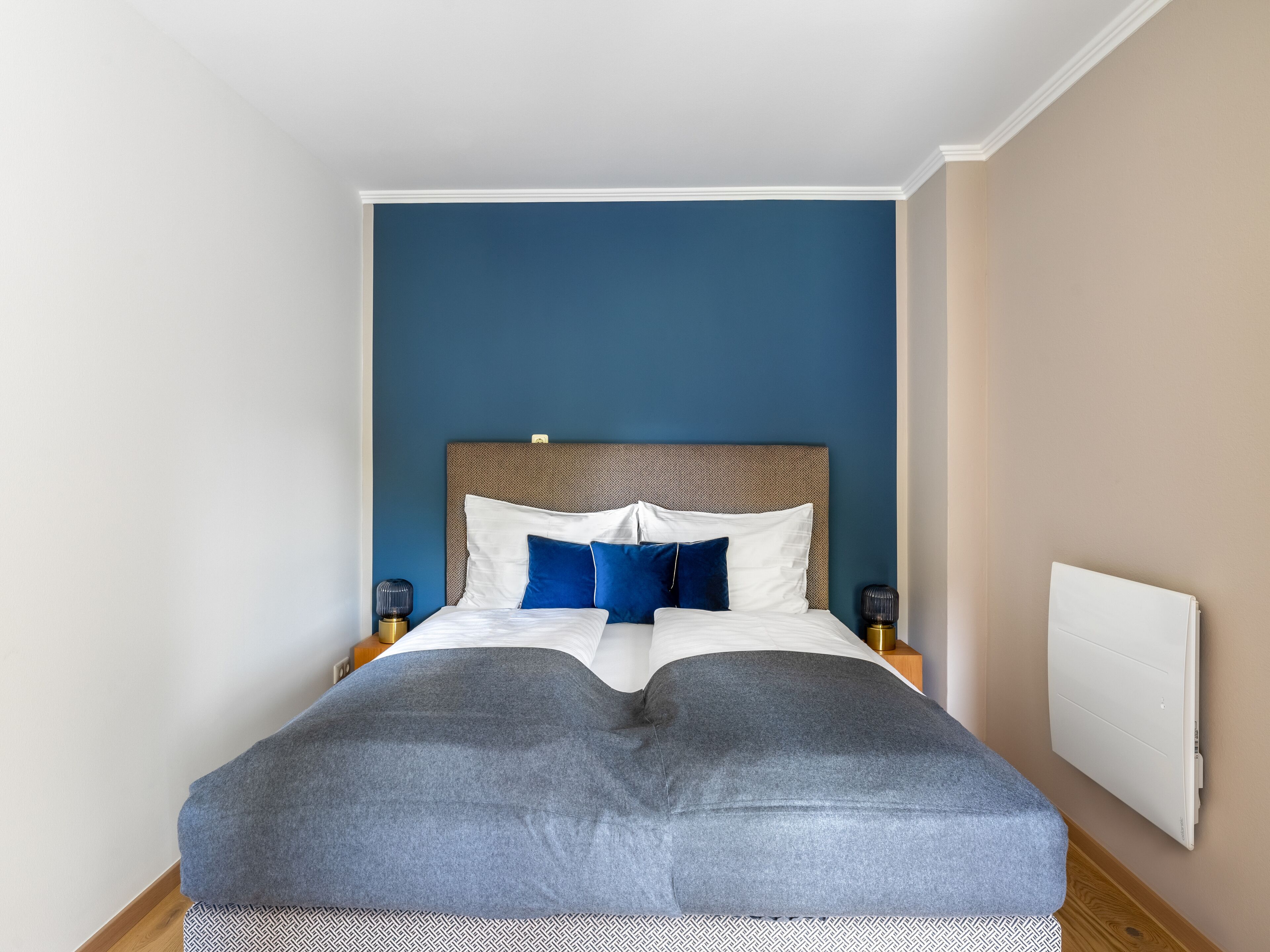 Ein ordentlich bezogenes Bett mit einem gemusterten Überwurf, zwei Nachttischlampen und einem Heizkörper an der Wand.