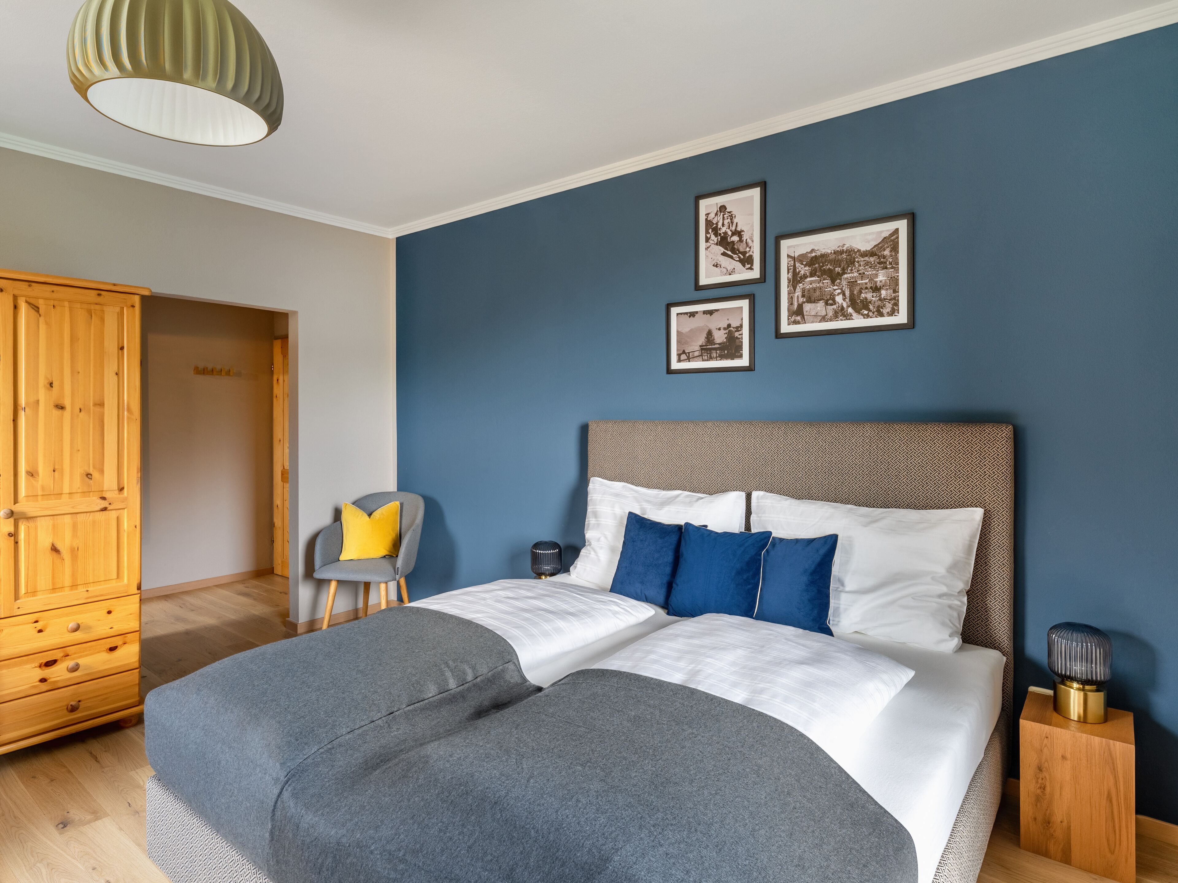 A bedroom with a bed, a wooden wardrobe, a chair, and two framed pictures on the wall.