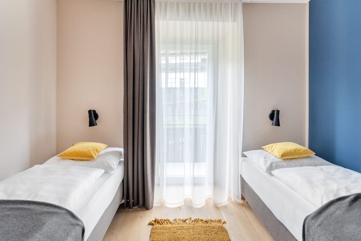 A hotel room with two beds, a large window with sheer curtains, a wooden floor, and a small rug.