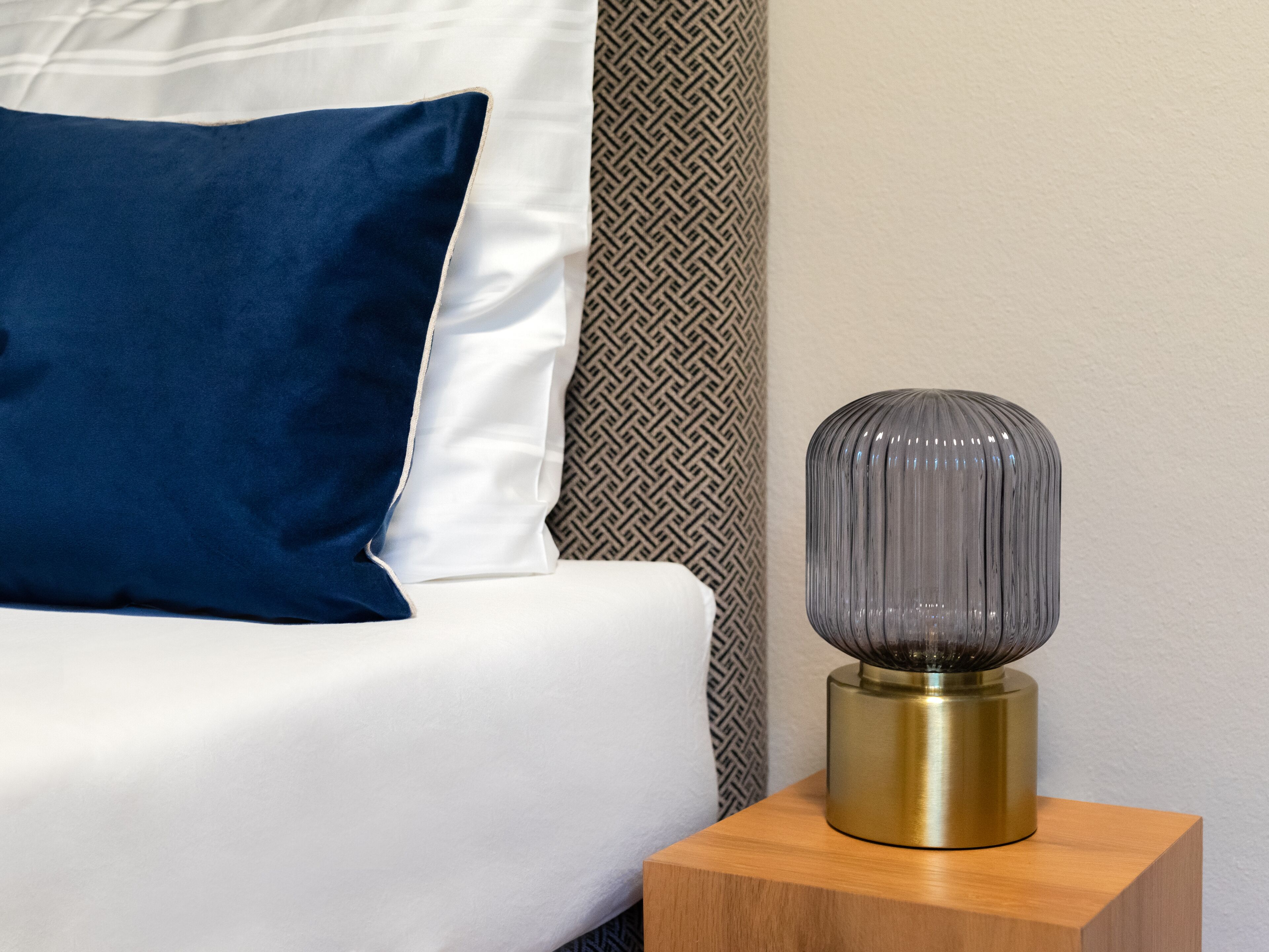 A bed with a white and blue pillow, a patterned wall, and a brass and glass lamp on a wooden nightstand.