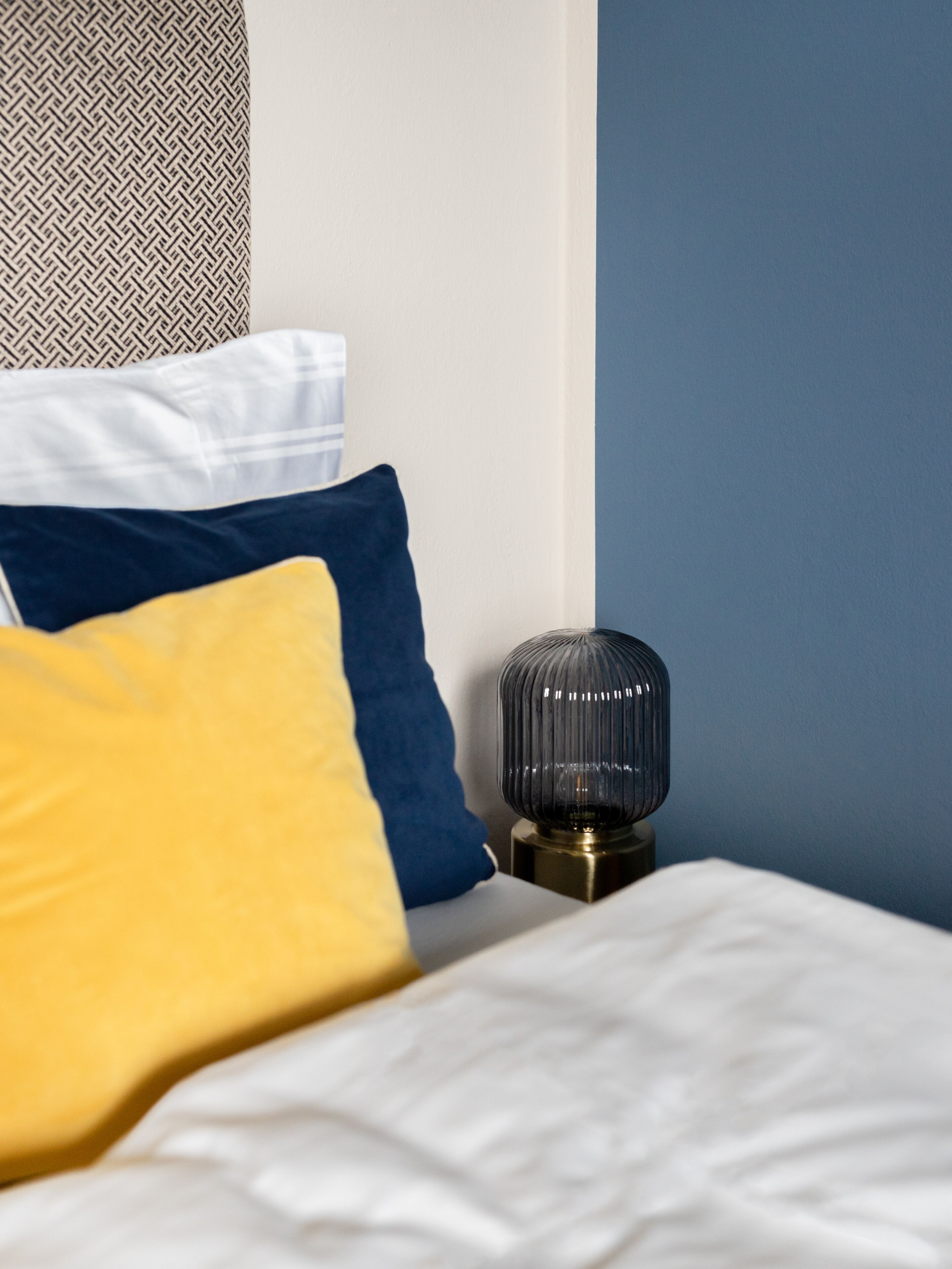 A neatly made bed with white and blue bedding, a decorative lamp, and patterned pillows.