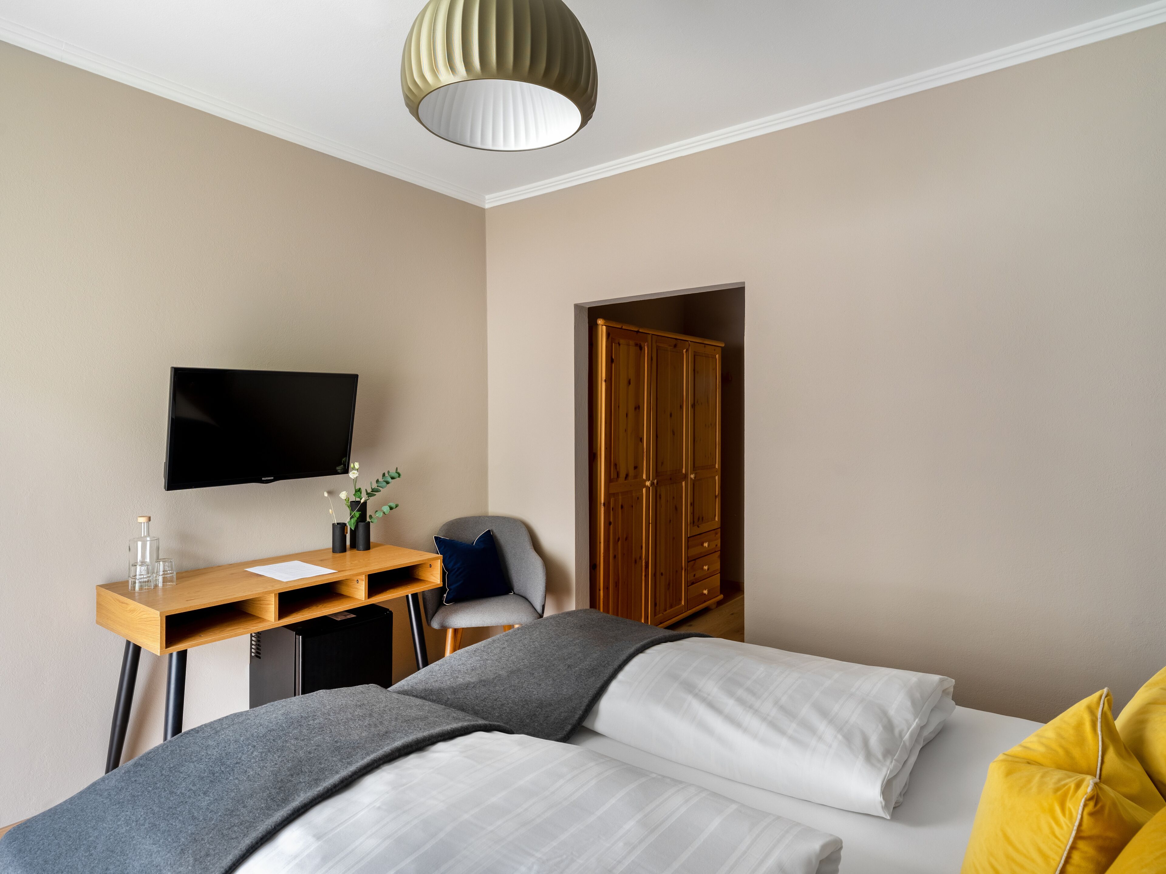 A hotel room with a bed, a desk with a flat-screen TV, a wardrobe, and a grey chair.