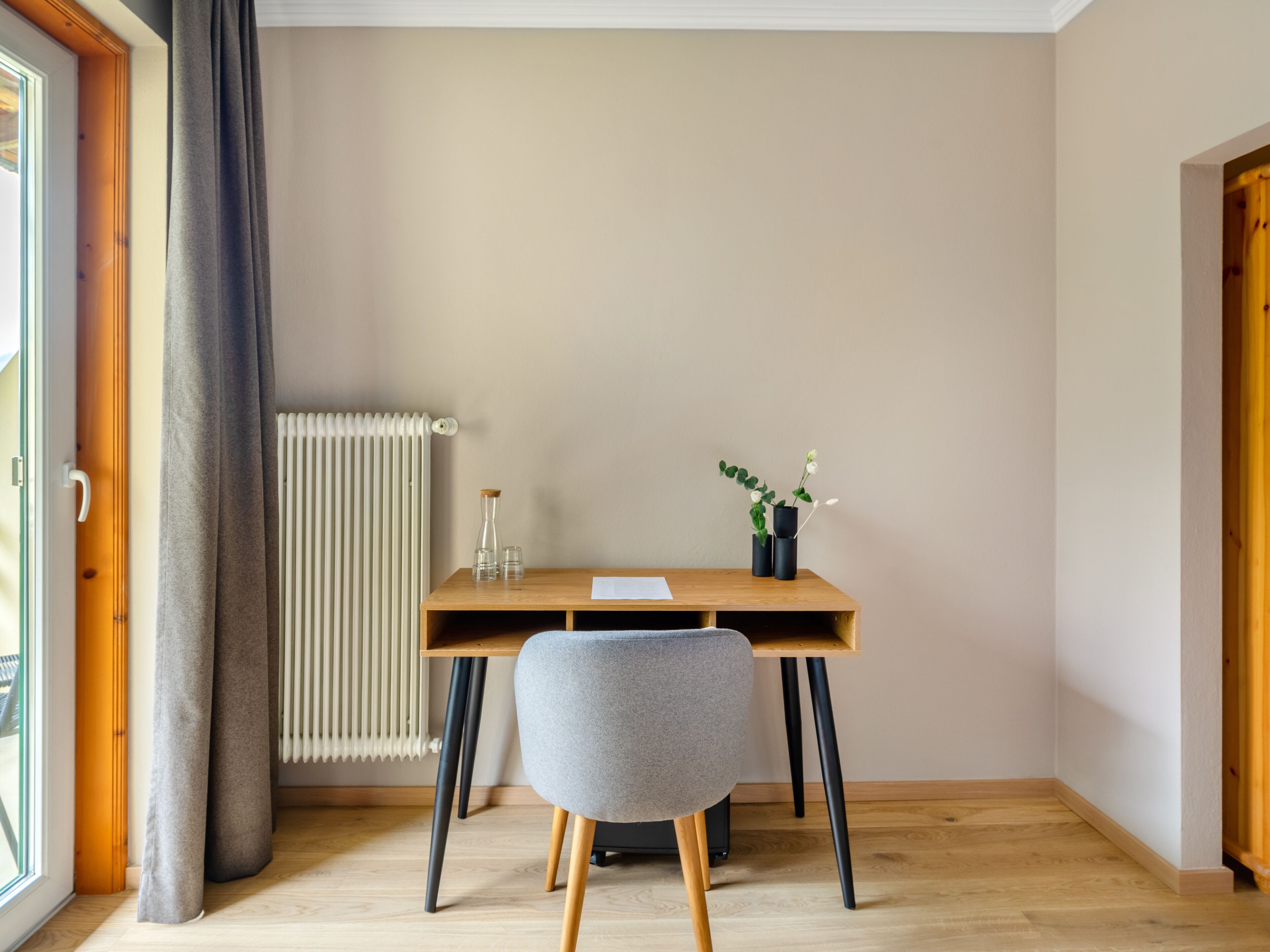 Une pièce avec un bureau en bois, une chaise grise, un radiateur et une fenêtre avec des rideaux.
