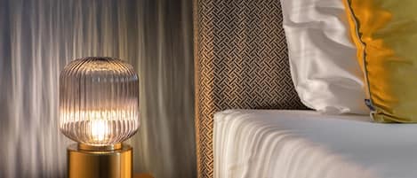A bed with white bedding and a gold pillow, a patterned wall, and a lamp with a ribbed base on a wooden nightstand.