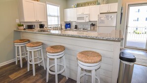 A kitchen with white cabinets, a granite countertop, and four bar stools with woven seats. - 3 night minimum.  2 bedroom. Outdoor pool. Close to beach. (North Myrtle Beach)