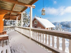 Uma varanda de madeira com guarda-corpo, um telhado coberto de neve e vista para uma casa ao fundo.