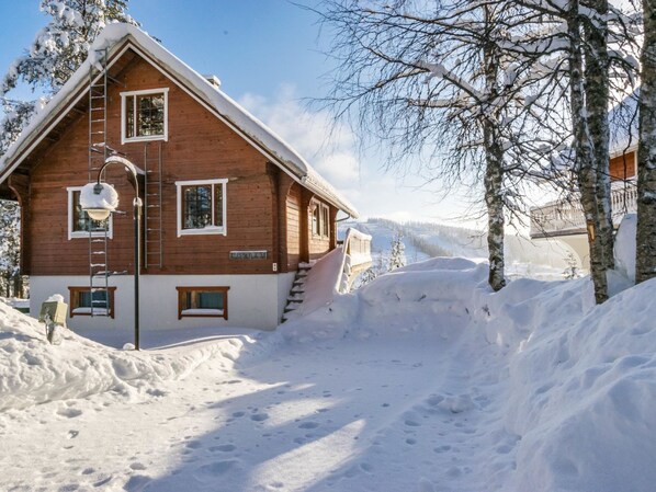 A wooden cabin with a snow-covered roof, a visible staircase, and a clear view of the surrounding snowy landscape.