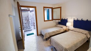 A bedroom with two single beds, a small table with a lamp, a mirror, and a view of a courtyard with a stone wall and a blue door. - Ca'Marin-Canal view and private patio (Venezia)