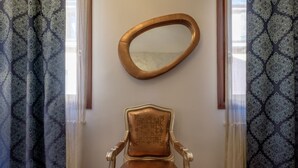 Quarto com cortina estampada, espelho oval fixado na parede e poltrona clássica com encosto decorativo.