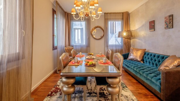 Uma área de refeições com uma mesa posta para quatro pessoas, rodeada por cadeiras, um lustre e um sofá com almofadas.