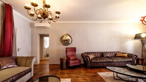 Uma sala de estar com um sofá de couro marrom, uma poltrona vermelha, uma mesa de centro de vidro e um lustre.